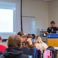 man behind a podium that has "Grand Valley State University" on a plaque; room full of college students listening